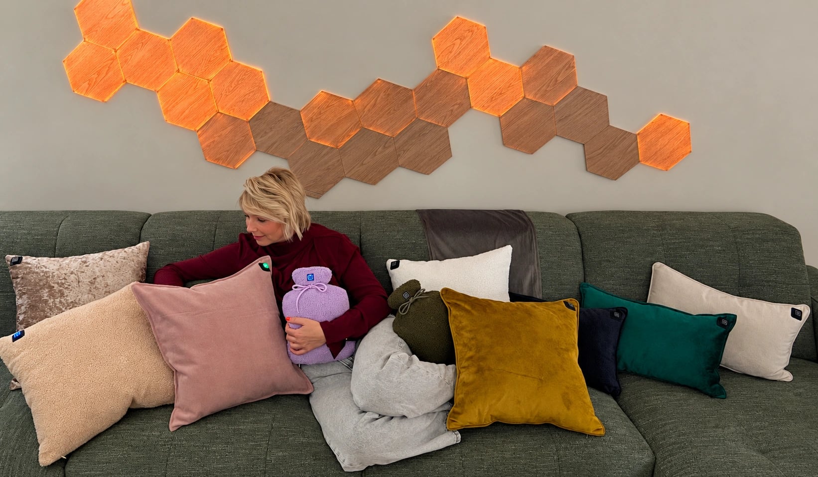 Une femme sur un canapé avec un coussin chauffant rose et un Warmpi Buddy violet, entourée de coussins de différentes couleurs et matières.